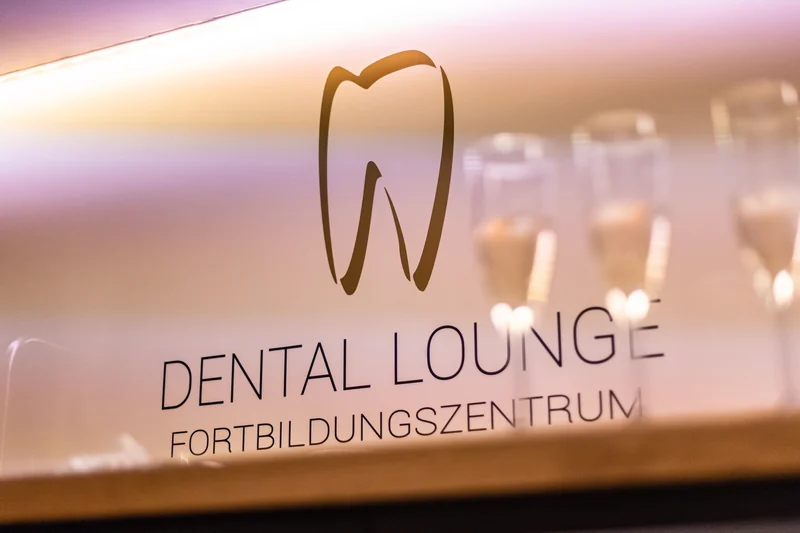 Golden beleuchtetes Dental-Lounge-Logo auf einer Glasfläche mit Sektflöten im Hintergrund